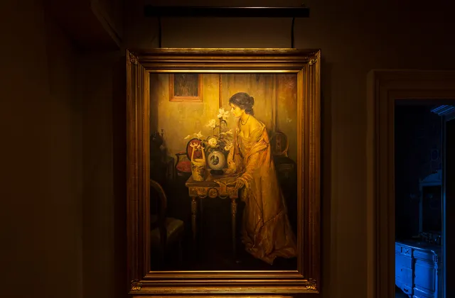 Painting of a lady leaning on a table with flowers in a vase lit by a picture light
