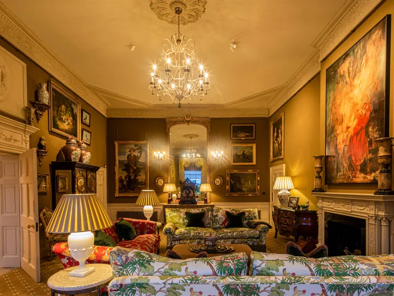 A rich drawing room in a country house wth central chandelier, architectural spotlights and decorative table lamps.