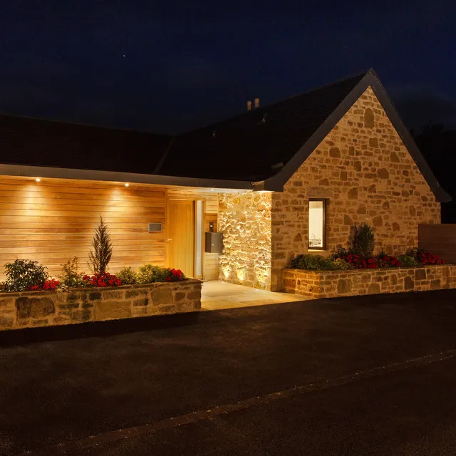 The front entrance of a new-build stone house at night.  Uplighting and downlighting picks out the textures of the stone and timber.