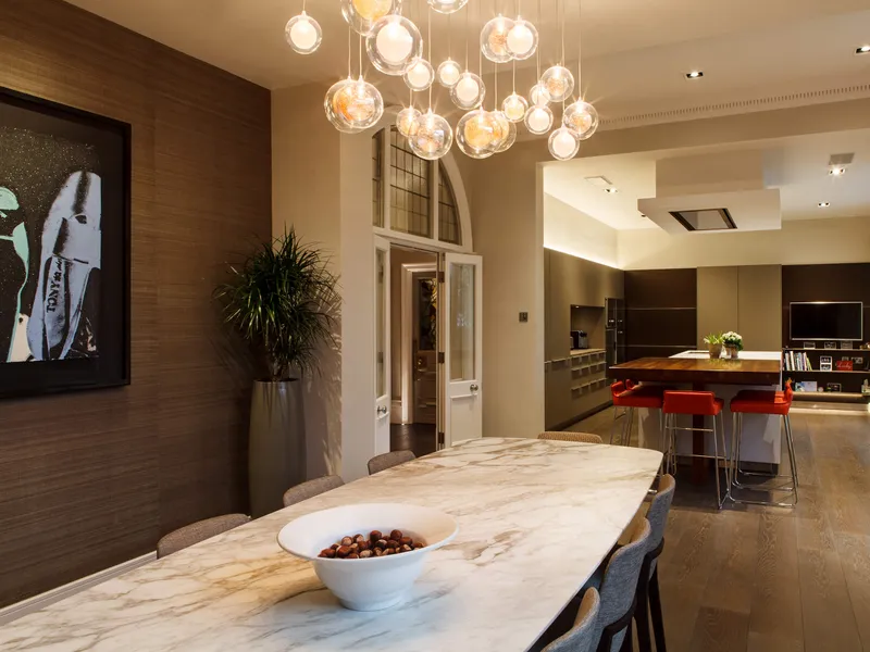 A sleek modern kitchen and dining area with strong architectural llighting and bespoke pendant over the dining table