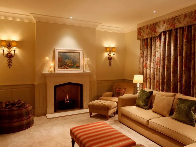 A drawing room in a country house with a fire and decorative wall and table lamps