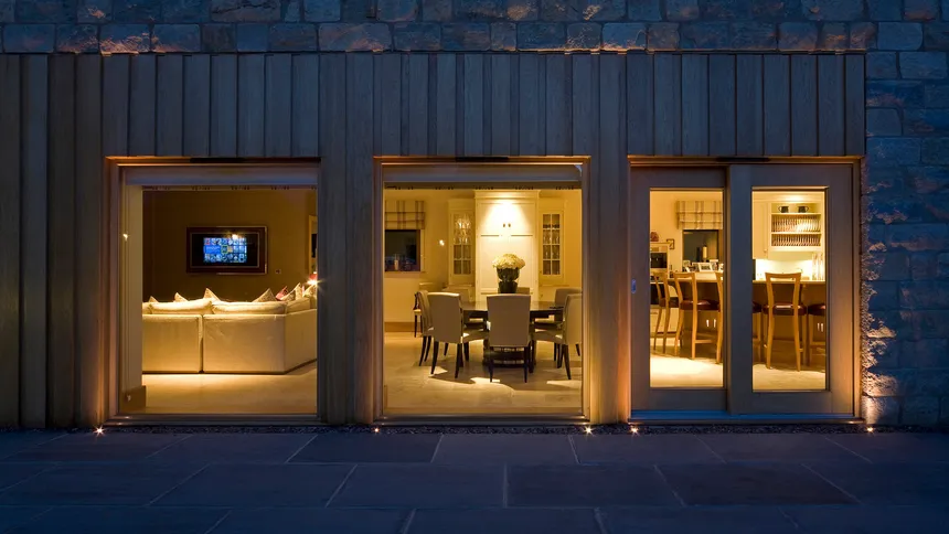 The exterior of a large new-build stone house at night looking in on a kitchen, dining area and TV lounge