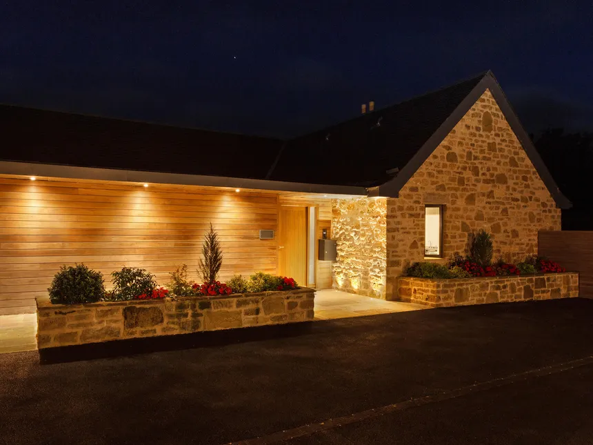 The front entrance of a new-build stone house at night.  Uplighting and downlighting picks out the textures of the stone and timber.